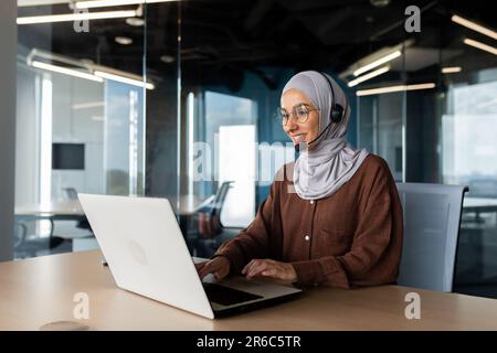 A young Muslim woman in a hijab and a headset is working in the office at a desk using a laptop. Typing on the keyboard, talking via video call, consulting. Stock Photo