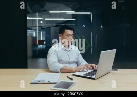 Serious and focused young asian businessman working overtime in office at desk behind laptop and documents. Stock Photo