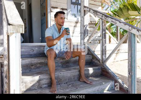 Thoughtful caucasian handsome young man drinking coffee while sitting on staircase outside cottage. Unaltered, vacation, tourist resort, lifestyle, dr Stock Photo