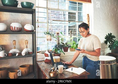 This customer wants something specific. an attractive young business owner standing and using her laptop and cellphone in her pottery studio. Stock Photo