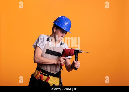 Female industrial engineer holding drilling gun in new interior renovation project, using power drill nail gun. Woman renovator wearing uniform in studio shot over yellow background. Stock Photo