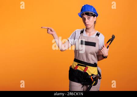 Woman builder indicating direction on left side of studio, posing with pair of pliers and pointing sideways. Female construction worker looking at camera and showing sides, yellow background. Stock Photo