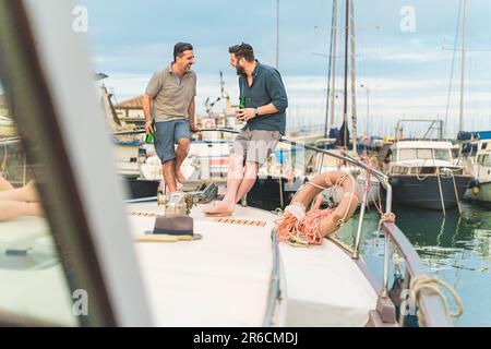Two caucasian friends in their forties having a conversation and smiling on the deck of a docked boat. They are dressed in casual summer attire, enjoy Stock Photo