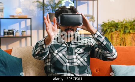 Indian man using virtual reality futuristic technology VR app headset helmet to play simulation 3D 360 video game, watching film movie at modern home apartment. Hindu guy in goggles sitting on couch Stock Photo