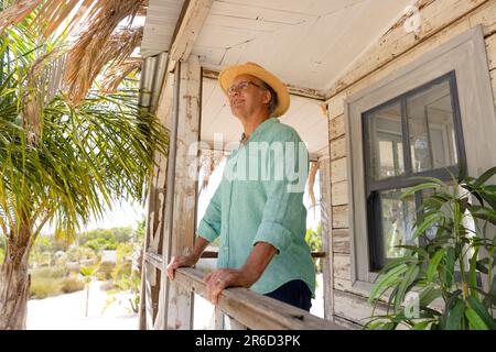 Caucasian senior man wearing hat smiling and contemplating while standing by railing in balcony Stock Photo