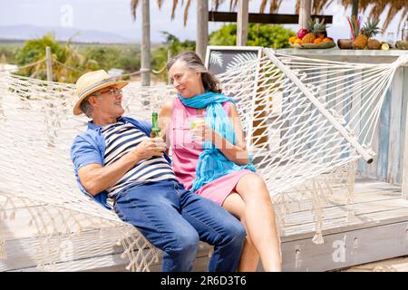 Caucasian senior couple holding drinks and talking while relaxing on hammock at beach Stock Photo