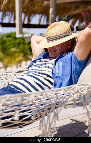 Caucasian senior man with hat on face and hands behind head sleeping on hammock at beach Stock Photo