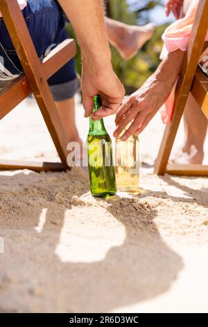 Cropped image of caucasian senior couple with beer bottles sitting on deck chairs at beach Stock Photo
