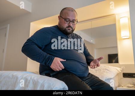 Huge man suffering from extra weight touching stomach sitting on bed in bedroom.  Stock Photo