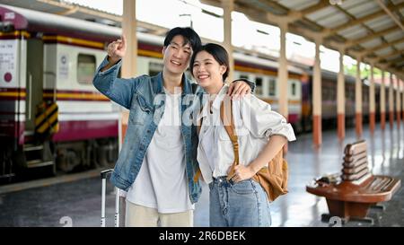 A happy and handsome young Asian man embraces his girlfriend while pointing his finger at something to show her while waiting to board the train at th Stock Photo