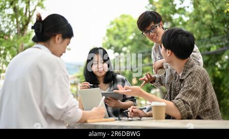 A group of smart and positive young Asian college students enjoy talking or sharing ideas on their project at a table in the campus park. Stock Photo