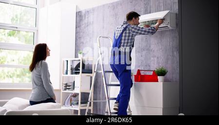 Smiling Woman Looking At Male Technician Repairing Air Conditioner With Screwdriver In Home Stock Photo