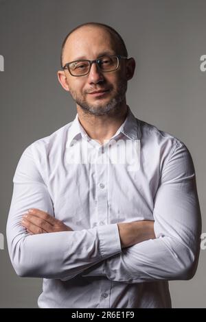 Relaxed portrait of a man in a white shirt and glasses with crossed arms and looking at the camera on a gray background Stock Photo