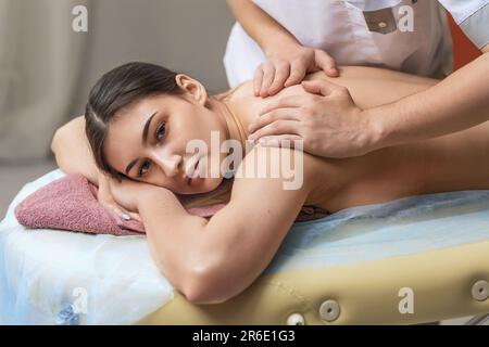 A male masseur makes a massage of the trapezius muscles and cervical region to a girl client in a treatment room, a young beautiful brunette in a spa Stock Photo