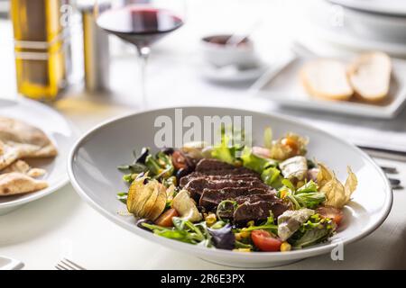 Beef steak sliced on top of mixed salad with greens, physalis, cherry tomatoes served with wine. Stock Photo