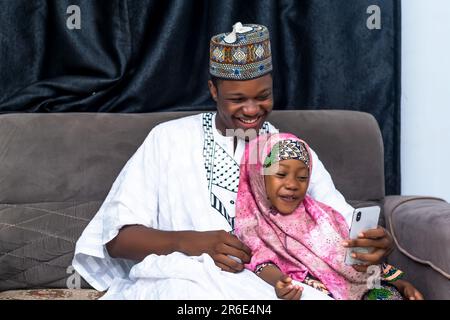 excited African dad and daughter seated viewing smartphone. Smiling, bonding and sharing positive emotions together. Stock Photo