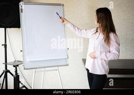 Concentrated young caucasian speaker woman coach show on white board, preparing or giving educational lecture. Female team leader explaining project i Stock Photo