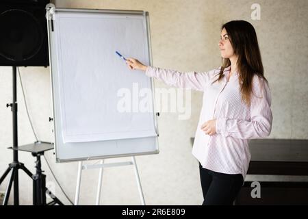 Concentrated young caucasian speaker woman coach show on white board, preparing or giving educational lecture. Female team leader explaining project i Stock Photo