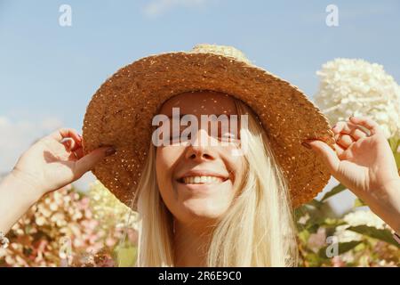 Happy young blonde woman in a straw hat near hydrangea flowers Stock Photo