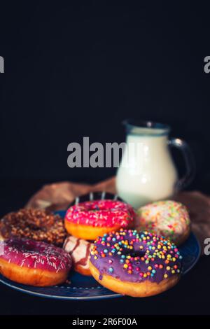 Assorted donuts in a plate and a jug of milk on a dark background. Stock Photo