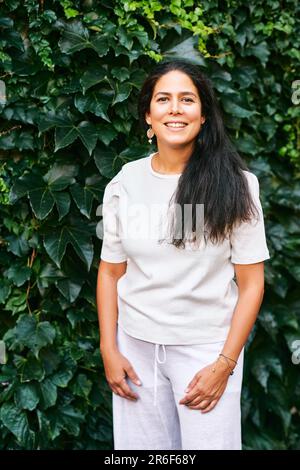 Outdoor portrait of cheerful middle age woman posing next to green ivy wall, wearing white clothes Stock Photo