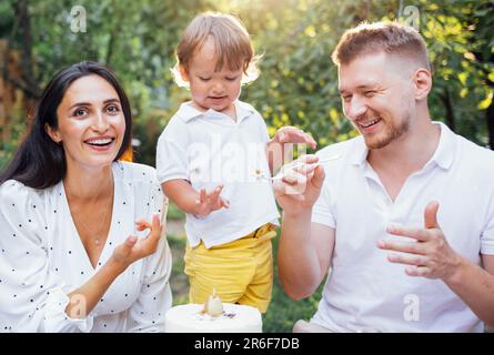 Little cute kid and his parents are tasting festive cake outdoors in garden. Happy family having fun. Smiling man holds out fork with piece of cake to Stock Photo