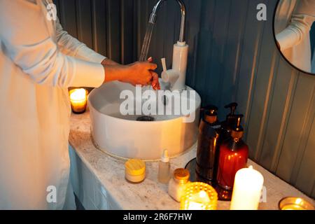 A young girl in white clothes washes her hands under running water over the sink. Cosmetics and detergents, lit scented candles on a marble tabletop. Stock Photo