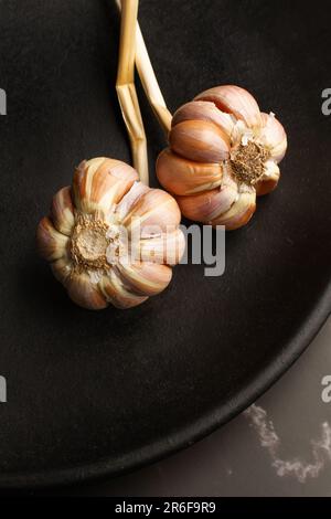 Two garlic heads on a black plate on a gray and white marble kitchen counter Stock Photo