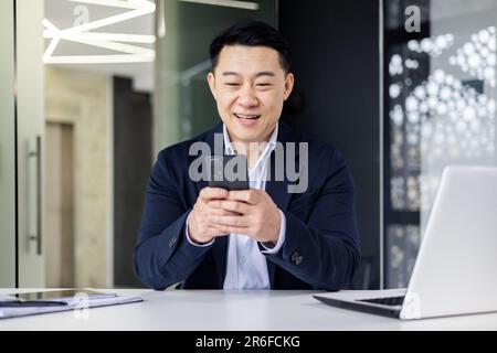 Young handsome Asian man, office worker sitting in the office at the table, fiddling with the phone, typing messages, chatting Stock Photo