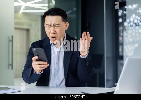 Confused Asian guy holding smart phone feels concerned thinking over received message. Mobile phone everyday usage, unpleasant news, waiting for important call, low signal device problems concept Stock Photo