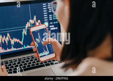 Close up of woman stock market investor analyst holding a smartphone with investment app. Female using laptop for cryptocurrency financial stock market analysis. Stock Photo