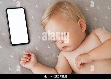 Closeup portrait of cute little boy watching sweet dreams in his bed falling asleep with music playing in app for baby sleep on phone with blank screen lying next to his head. Maternity and technology Stock Photo