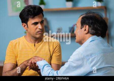 Shoulder shot, indian adult son consoling his senior father by holding hands while sitting on sofa at home - concept of family support, relationship Stock Photo