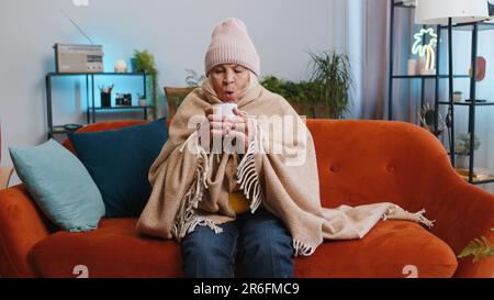 Sick senior grandmother woman in plaid shivering from cold on sofa drinking hot tea in unheated apartment without heating due debt. Unhealthy ill elderly pensioner feeling discomfort try to warming up Stock Photo