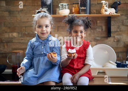 Two happy girls sitting in kitchen countertop, enjoying muffins together getting messy with food, feeling joyful. Home, family, lifestyle concept. Stock Photo