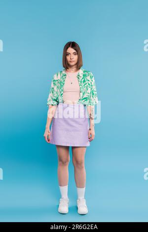 gen z fashion, young brunette woman with short hair posing in shirt with palm tree print, skirt and white sneakers on blue background, carefree, tatto Stock Photo