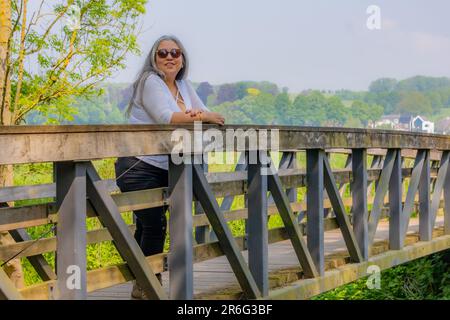 Mature brunette woman leaning on fence of bridge, trees in misty background, looking at camera, white blouse, sunglasses, long gray hair, sunny day in Stock Photo