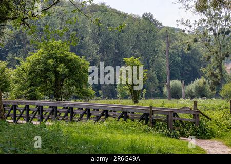 Valley with a wooden bridge over Geul river in Dutch nature park Ingendael, mountain covered with lush trees in background, sunny spring day on hiking Stock Photo