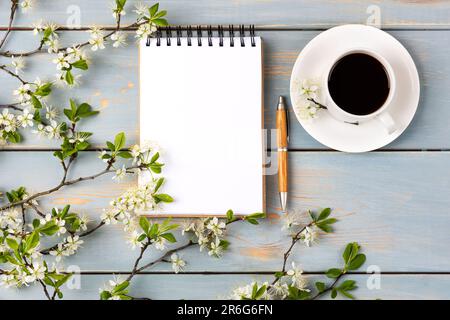Artistic composition with opened blank notepad and pen, cup of black coffee, branch of white cherry blossoms on blue wooden table. Festive office desk Stock Photo
