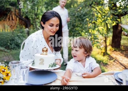 Little cute kid and his parents are tasting festive cake outdoors in garden. Happy family having fun. Smiling man holds out fork with piece of cake to Stock Photo