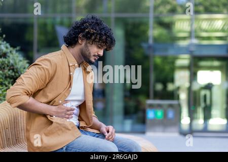 A young Hispanic man is sitting on a bench outside and holding his stomach with his hands. Suffers from pain, discomfort, feels bad. Stock Photo