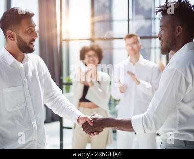 Glad to work with you. Young modern men in smart casual wear shaking hands and smiling while working in the creative office Stock Photo