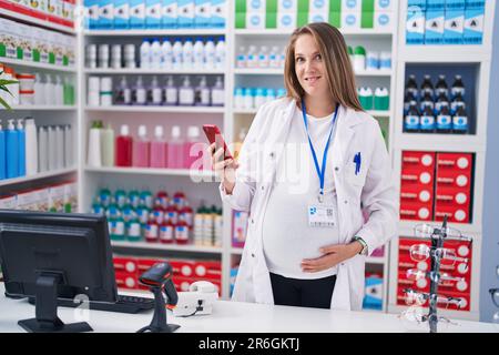 Young pregnant woman pharmacist using smartphone working at pharmacy Stock Photo