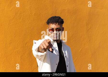 A male adult wearing sunglasses making a gesture of pointing with his finger towards the camera, against a bright orange background Stock Photo