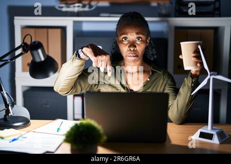 African woman working using computer laptop at night pointing down looking sad and upset, indicating direction with fingers, unhappy and depressed. Stock Photo