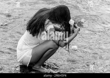 A high-contrast black and white image of a woman kneeling in shallow water, her hands cupping several small pebbles Stock Photo