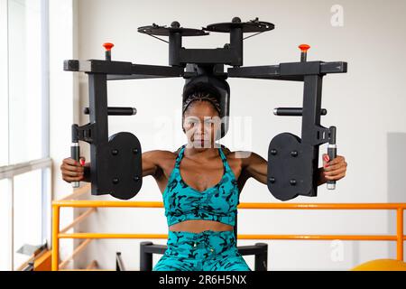 A young female is standing at a gym machine, using two dumbbells to perform an exercise Stock Photo