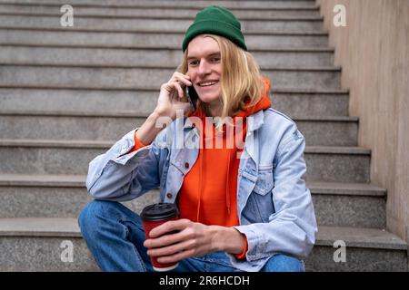 Young hapyp guy talk on phone and sitting on staircase in street, drinking coffee in between work Stock Photo