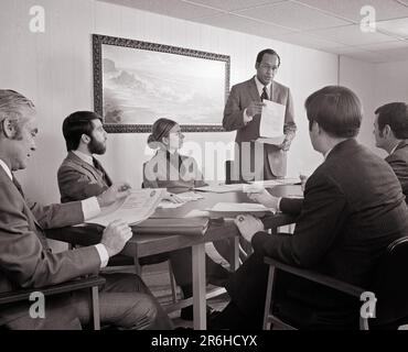 1970s AFRICAN-AMERICAN MAN STANDING READING A REPORT TO A GROUP MEN AND A WOMAN SITTING AROUND CONFERENCE TABLE  - o3083 HAR001 HARS COMMUNICATION TEAMWORK INFORMATION LIFESTYLE FIVE FEMALES 5 JOBS 6 MANAGER COPY SPACE REPORT HALF-LENGTH LADIES PERSONS MALES SIX CORPORATE CONFIDENCE EXECUTIVES MIDDLE-AGED B&W BUSINESSWOMAN MIDDLE-AGED MAN GOALS SKILL OCCUPATION SKILLS DISCOVERY AFRICAN-AMERICANS AFRICAN-AMERICAN AND LEADERSHIP BLACK ETHNICITY OPPORTUNITY OCCUPATIONS BOSSES SUPPORT BOARD ROOM BUSINESSWOMEN CONFERENCE ROOM COOPERATION MANAGERS MID-ADULT MID-ADULT MAN MID-ADULT WOMAN SOLUTIONS Stock Photo