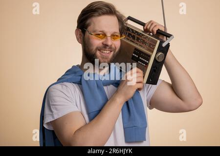 Happy Caucasian man using retro tape record player to listen music, disco dancing of favorite track, having fun, entertaining, fan of vintage technologies. Handsome guy isolated on beige background Stock Photo